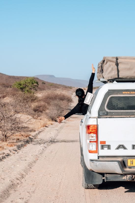 A passenger extends their arm out the window of a white 4x4 vehicle driving along a dry dirt road in Namibia.