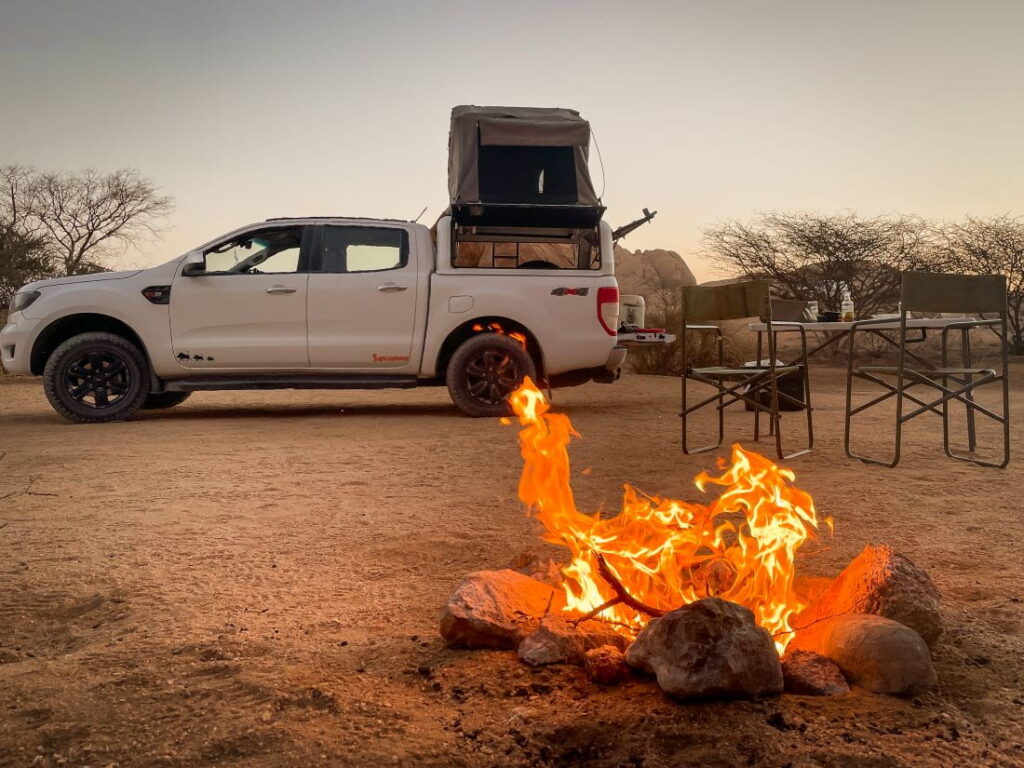 A white 4x4 truck with a pitched rooftop tent parked next to a glowing campfire at a desert campsite in Namibia.