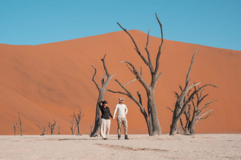 Matthias and Mieken holding hands in Deadvlei, standing beside ancient dead trees with a massive orange sand dune behind them.