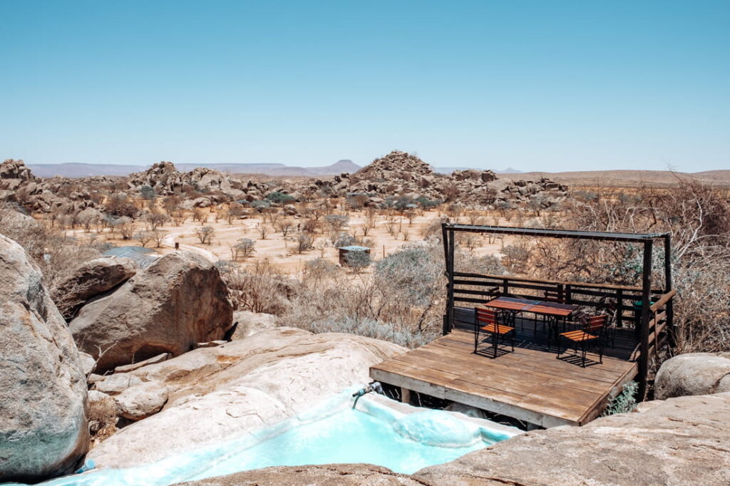 A wooden deck with a patio set overlooks a small pool built into natural granite boulders in a dry valley.