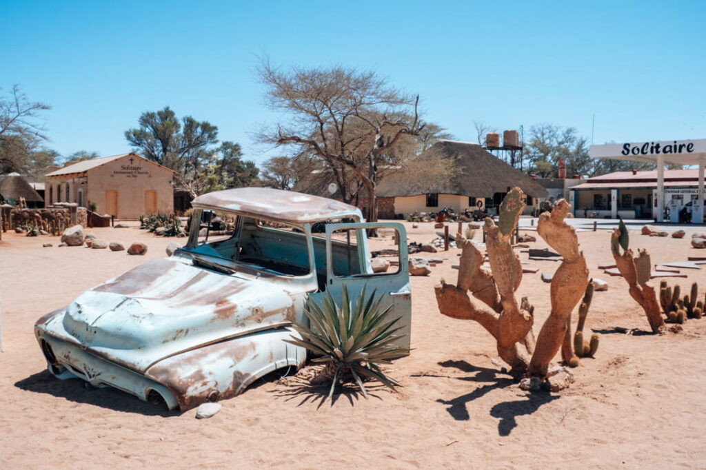 A rusted antique car shell is buried in the sand next to large cacti in Solitaire, Namibia.