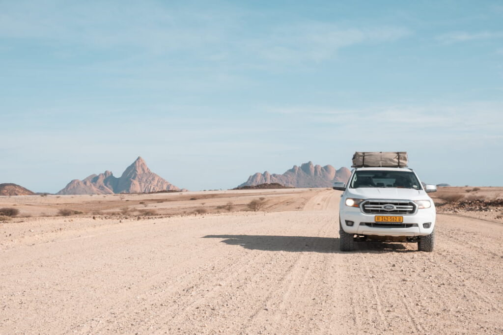A white 4x4 pickup truck with a rooftop tent is parked on a dirt road facing distant mountains.