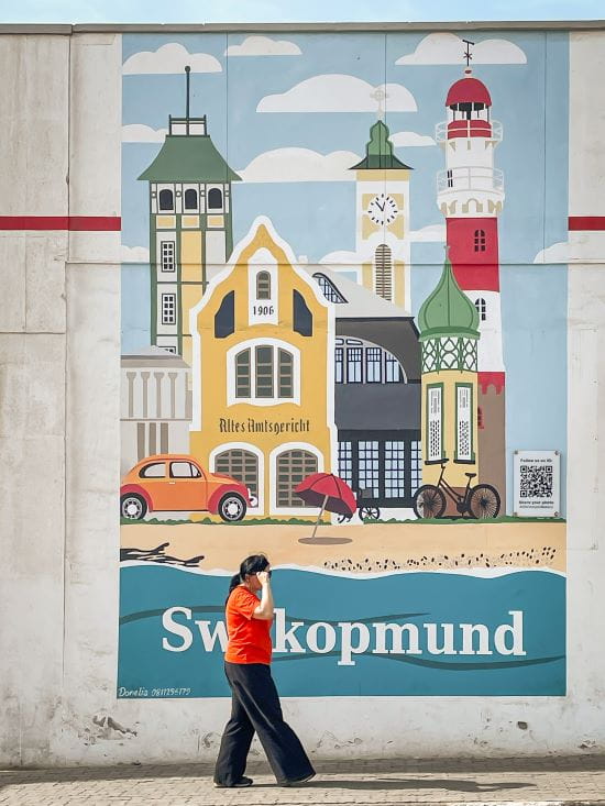 Mieke walking past a colorful mural depicting iconic landmarks and architecture of Swakopmund.