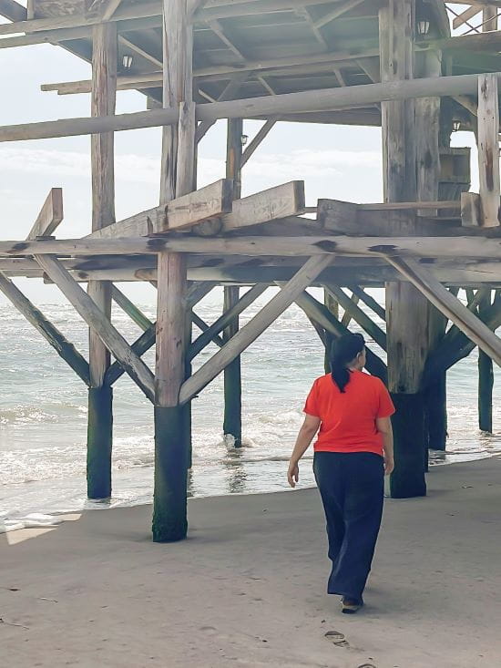 Mieke wearing a red shirt walks under the large wooden supports of the Swakopmund Jetty over the sand.