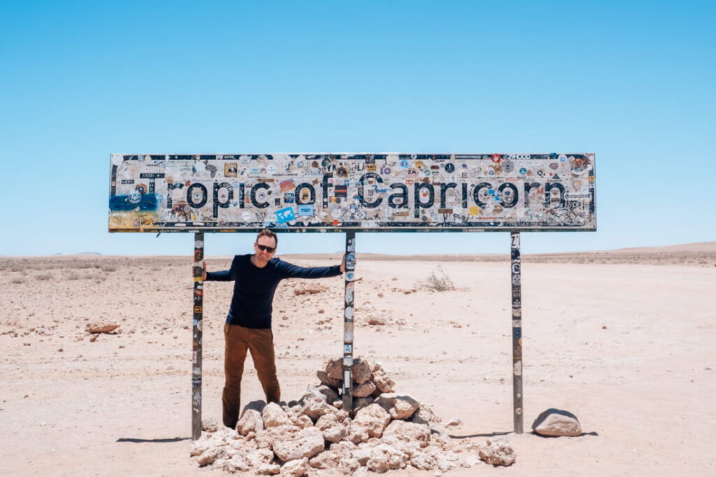 Matthias is smiling under the sticker-covered Tropic of Capricorn road sign in a flat desert.