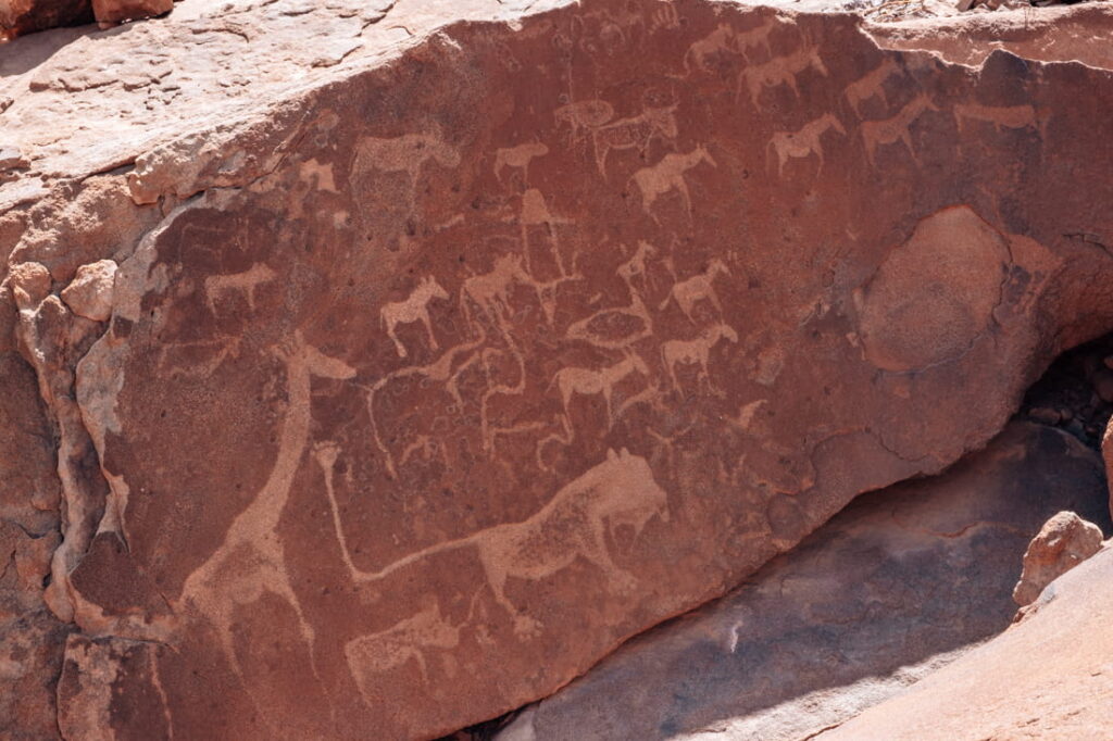 Ancient San rock engravings of a giraffe, a lion, and other animals carved into red sandstone.
