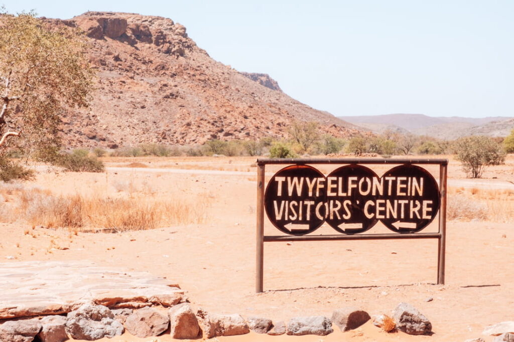 A dark metal sign for the Twyfelfontein Visitors' Centre stands in front of a rocky desert mountain.