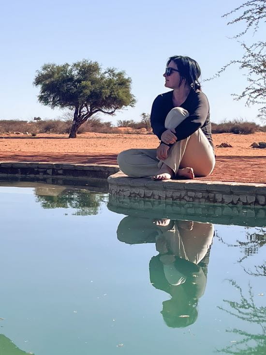 Mieke zit met opgetrokken knieën aan de rand van een zwembad in een droog woestijnlandschap met haar weerspiegeling in het water.