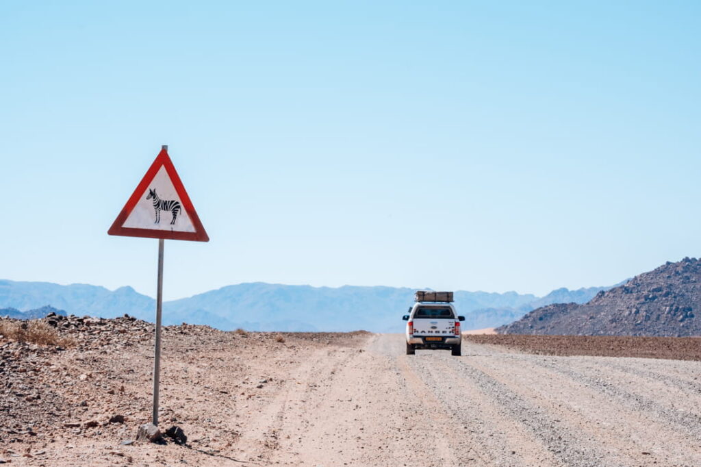 Een witte 4x4 pick-up met een daktent rijdt op een brede gravelweg langs een waarschuwingsbord met een zebra, tegen een achtergrond van blauwe bergen.