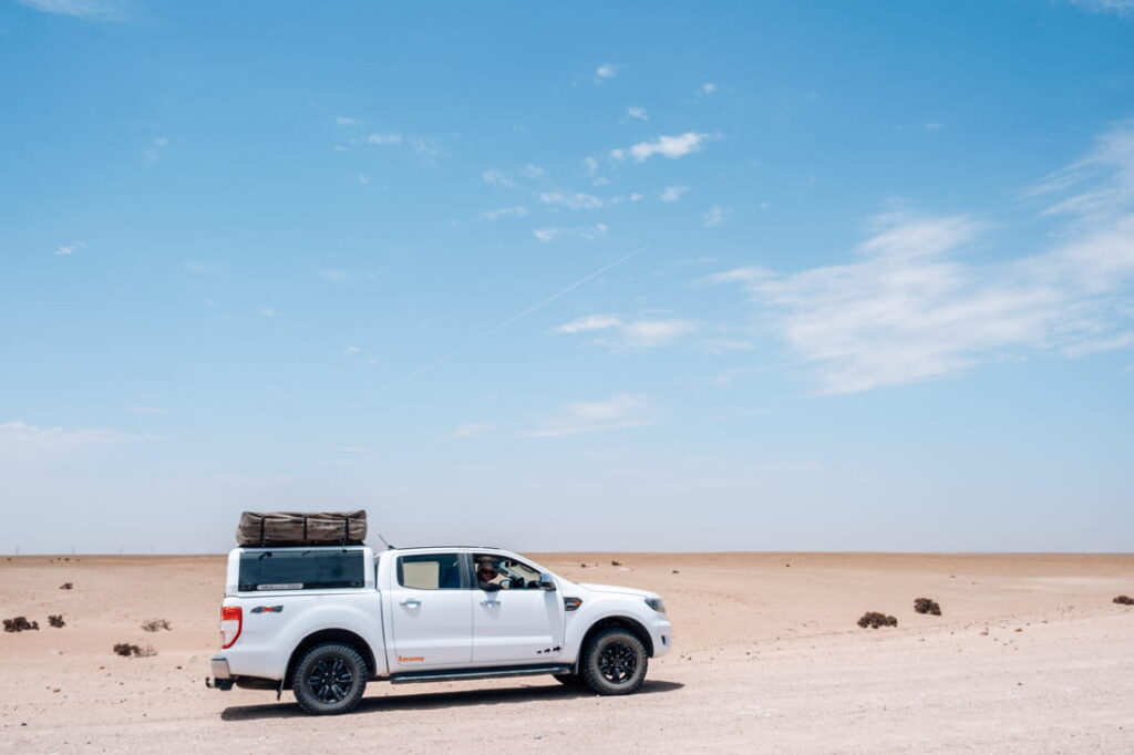 White 4x4 vehicle equipped with a rooftop tent driving along a gravel road in the Namib Desert, Namibia.