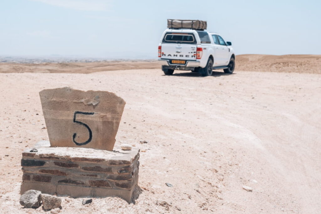 Numbered stone beacon marker 5 along a desert gravel track with a white pickup truck parked in the background.