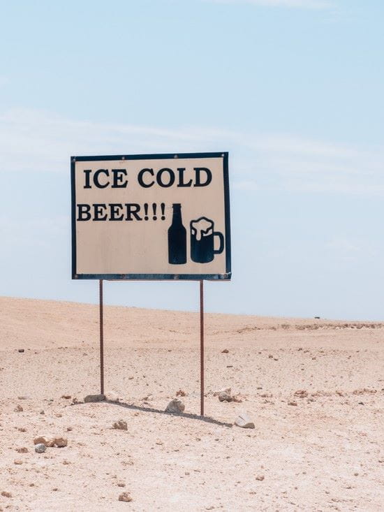 White sign with black text reading ICE COLD BEER standing in the sandy Namib Desert, Namibia.