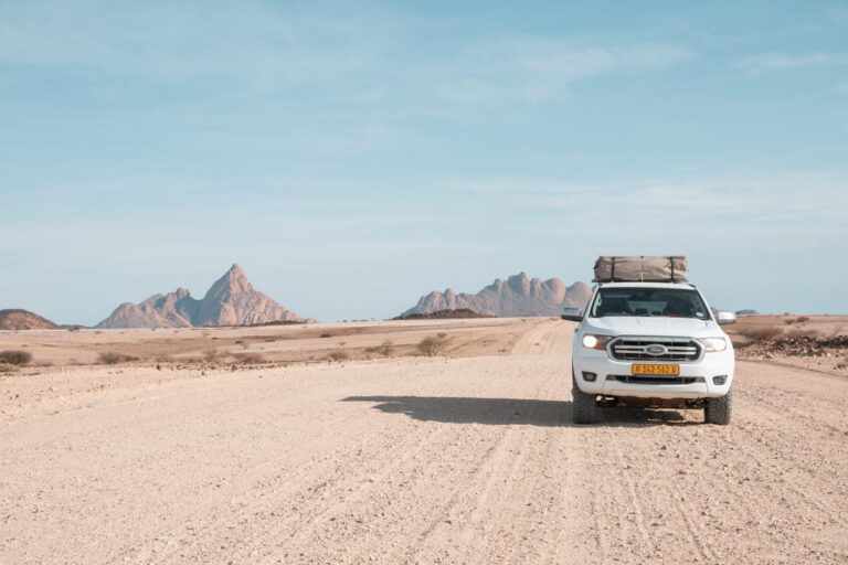 A white 4x4 truck with a rooftop tent on a dirt road facing the Spitzkoppe granite peaks in Namibia.